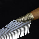 An extreme close-up of a custom fixed knife, highlighting the transition between the textured ridged brass bolster and the engraved silver feather blade.