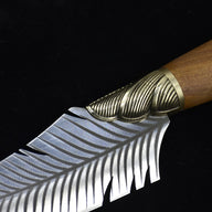 An extreme close-up of a custom fixed knife, highlighting the transition between the textured ridged brass bolster and the engraved silver feather blade.