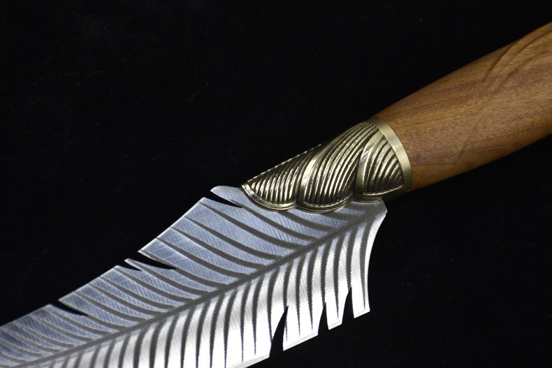An extreme close-up of a custom fixed knife, highlighting the transition between the textured ridged brass bolster and the engraved silver feather blade.
