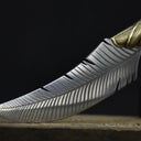 A close-up of the curved tip and textured edge of a custom silver feather blade knife, resting on an old wooden surface.