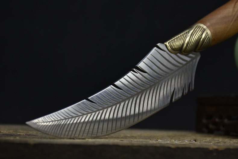 A close-up of the curved tip and textured edge of a custom silver feather blade knife, resting on an old wooden surface.