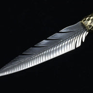 A diagonal view of a handmade fixed blade knife, highlighting the transition from the hammered brass bolster to the unique feather-patterned silver blade.