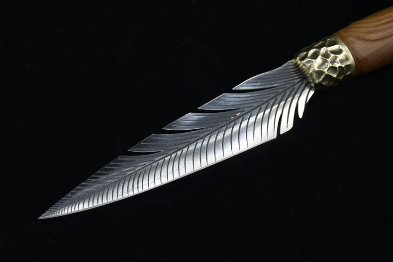 A diagonal view of a handmade fixed blade knife, highlighting the transition from the hammered brass bolster to the unique feather-patterned silver blade.