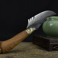 A beautifully crafted curved feather blade knife with a wooden handle, displayed in front of a green celadon jar on a rustic wooden surface.