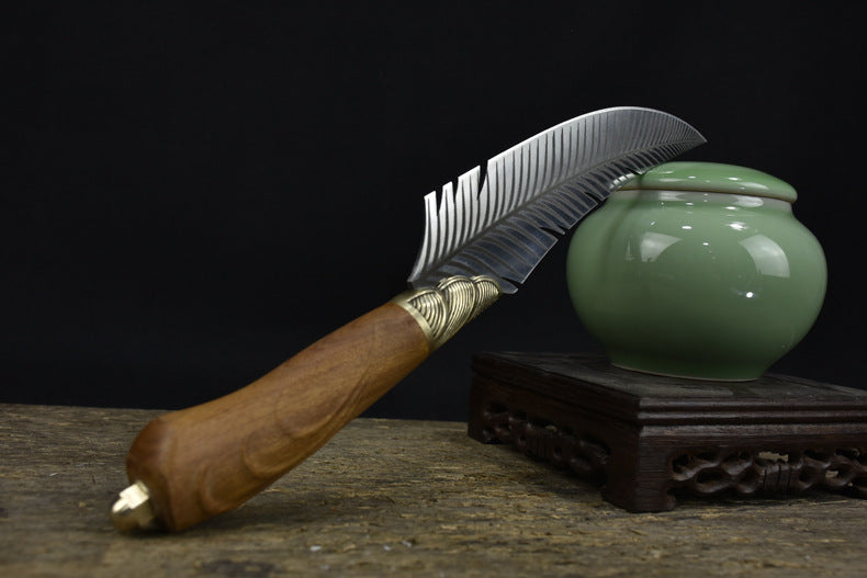 A beautifully crafted curved feather blade knife with a wooden handle, displayed in front of a green celadon jar on a rustic wooden surface.