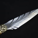 A close-up view of a custom fixed blade knife, capturing the light reflections on the intricate feather-patterned silver blade and the textured brass bolster.