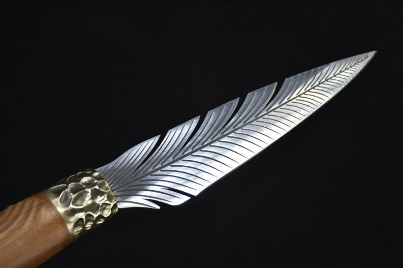A close-up view of a custom fixed blade knife, capturing the light reflections on the intricate feather-patterned silver blade and the textured brass bolster.