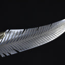 A close-up of a custom fixed blade knife showing intricate feather engraving on the silver blade and a ridged brass bolster, displayed on a dark background.