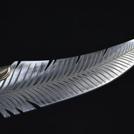 A close-up of a custom fixed blade knife showing intricate feather engraving on the silver blade and a ridged brass bolster, displayed on a dark background.