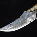 A close-up of the curved silver blade of a fixed knife, featuring detailed feather engraving and a ridged brass bolster, displayed on a dark background.