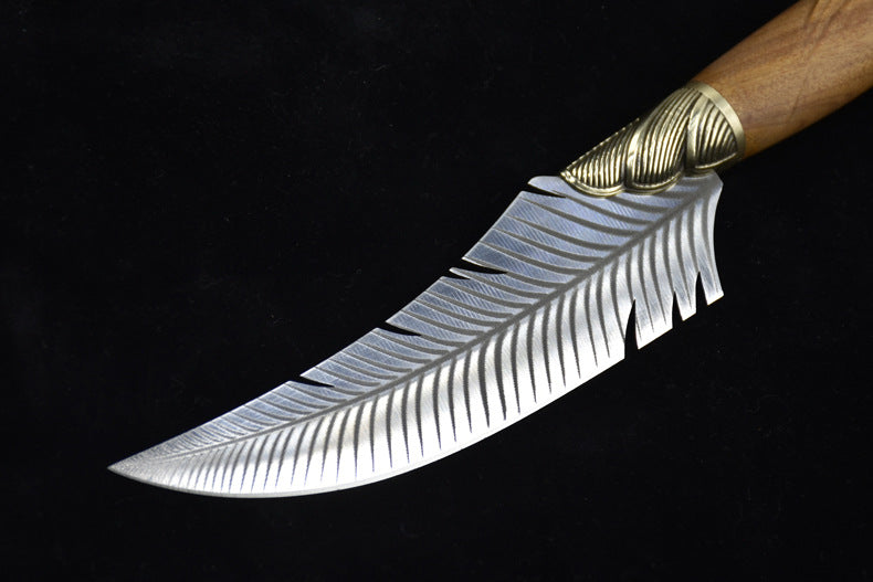 A close-up of the curved silver blade of a fixed knife, featuring detailed feather engraving and a ridged brass bolster, displayed on a dark background.