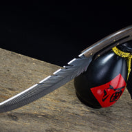 A full side view of an engraved feather blade knife resting on a wooden surface, displayed next to a black liquor gourd with a red label and yellow rope.