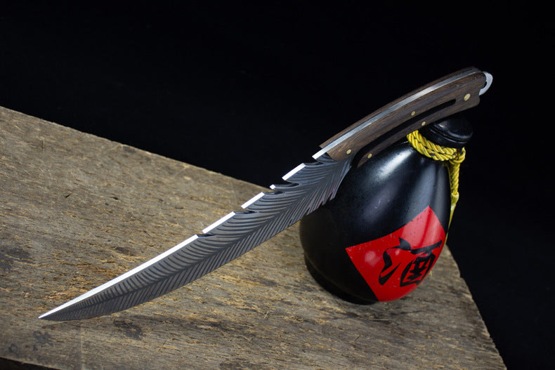 A full side view of an engraved feather blade knife resting on a wooden surface, displayed next to a black liquor gourd with a red label and yellow rope.