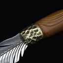 An extreme close-up of a custom fixed blade knife, showcasing the hammered brass bolster, grooved wooden handle, and intricate silver feather-engraved blade.