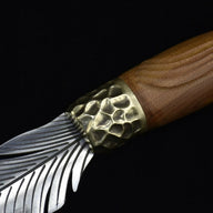 An extreme close-up of a custom fixed blade knife, showcasing the hammered brass bolster, grooved wooden handle, and intricate silver feather-engraved blade.