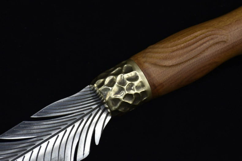 An extreme close-up of a custom fixed blade knife, showcasing the hammered brass bolster, grooved wooden handle, and intricate silver feather-engraved blade.