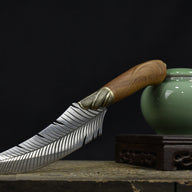 An engraved feather blade knife with a wooden handle resting its handle on a green celadon jar, displaying the full curved profile against a dark background.