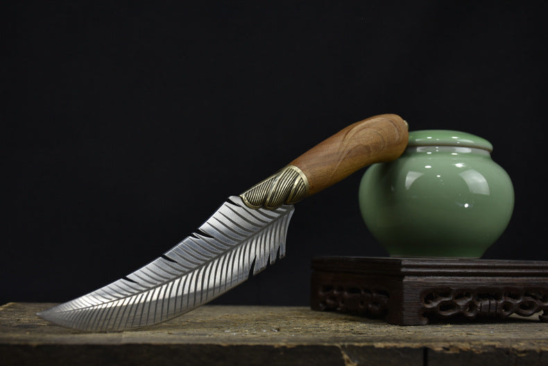 An engraved feather blade knife with a wooden handle resting its handle on a green celadon jar, displaying the full curved profile against a dark background.