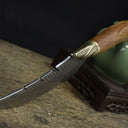 A side view of a custom fixed blade knife resting on a textured wooden board, highlighting the curved blade, brass bolster, and wooden handle.