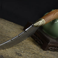 A side view of a custom fixed blade knife resting on a textured wooden board, highlighting the curved blade, brass bolster, and wooden handle.