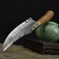 A fixed blade knife with a feather-engraved blade and a wooden handle, displayed diagonally next to a small green ceramic jar on a wooden stand.