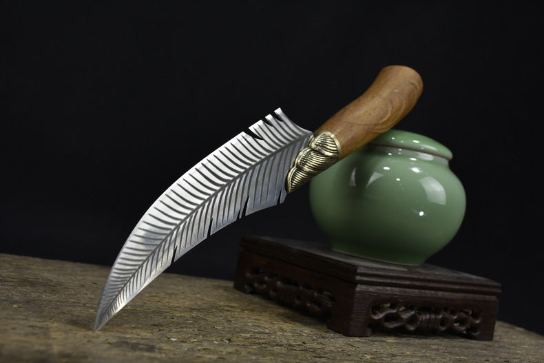 A fixed blade knife with a feather-engraved blade and a wooden handle, displayed diagonally next to a small green ceramic jar on a wooden stand.