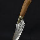 An artisan fixed knife with a feather-engraved blade and a wooden handle standing at an angle on a wooden surface against a dark background.