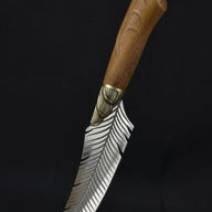 An artisan fixed knife with a feather-engraved blade and a wooden handle standing at an angle on a wooden surface against a dark background.