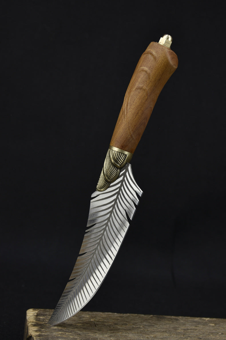 An artisan fixed knife with a feather-engraved blade and a wooden handle standing at an angle on a wooden surface against a dark background.