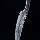An engraved feather blade knife standing vertically on a wooden surface with its tip pointing downwards, displaying the handle and blade in a dark background.