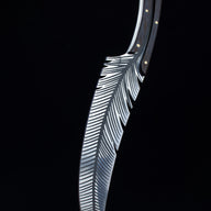 An engraved feather blade knife standing vertically on a wooden surface with its tip pointing downwards, displaying the handle and blade in a dark background.