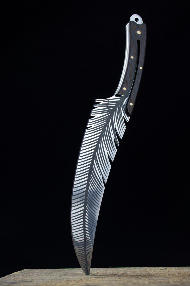 An engraved feather blade knife standing vertically on a wooden surface with its tip pointing downwards, displaying the handle and blade in a dark background.