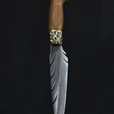 A handmade fixed blade knife with a grooved wooden handle and a hammered brass bolster, standing vertically on a dark surface.