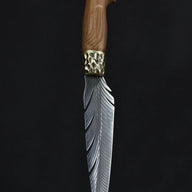 A handmade fixed blade knife with a grooved wooden handle and a hammered brass bolster, standing vertically on a dark surface.