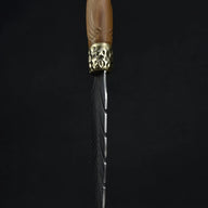 An artisan fixed blade knife standing vertically on a wooden surface, showing its elegant wooden handle and feather-engraved blade against a dark background.