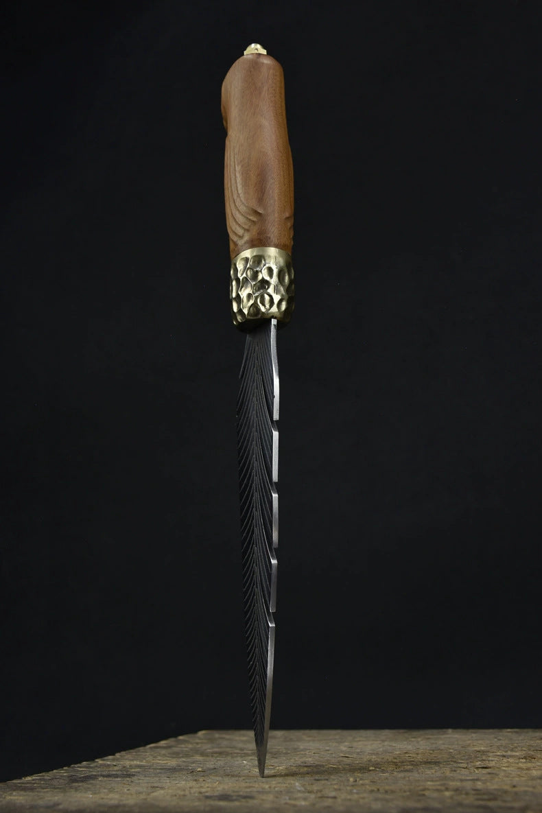 An artisan fixed blade knife standing vertically on a wooden surface, showing its elegant wooden handle and feather-engraved blade against a dark background.