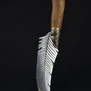 A handcrafted curved feather blade knife standing vertically on a wooden surface with its tip pointing downwards, displaying the handle and brass bolster against a dark background.