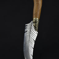 A handcrafted curved feather blade knife standing vertically on a wooden surface with its tip pointing downwards, displaying the handle and brass bolster against a dark background.