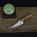 A top-down view of a curved feather blade knife with a wooden handle, placed next to a green celadon jar on a dark wooden board against a black background.
