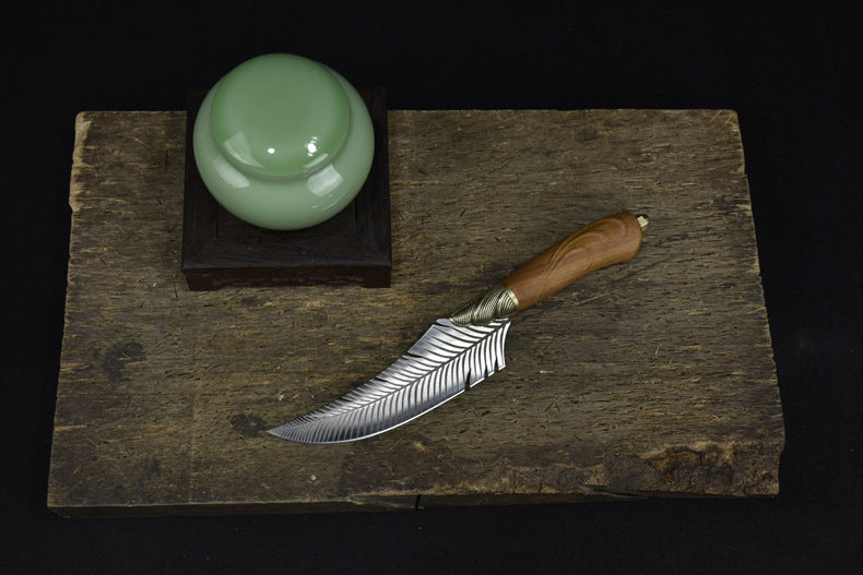 A top-down view of a curved feather blade knife with a wooden handle, placed next to a green celadon jar on a dark wooden board against a black background.