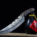 An artistic shot of the custom feather blade knife leaning against a black liquor gourd with a red label on a dark surface.