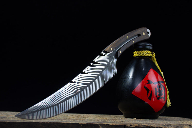 An artistic shot of the custom feather blade knife leaning against a black liquor gourd with a red label on a dark surface.