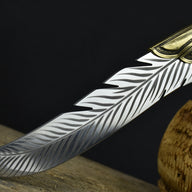 A close-up of the tip of an engraved silver feather blade, showcasing the curved shape and detailed feather engraving.