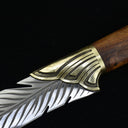 A close-up showing the transition between the textured brass bolster and the engraved silver feather blade of a fixed knife.