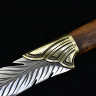 A close-up showing the transition between the textured brass bolster and the engraved silver feather blade of a fixed knife.