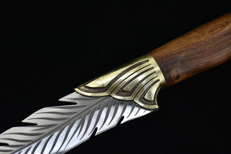 A close-up showing the transition between the textured brass bolster and the engraved silver feather blade of a fixed knife.