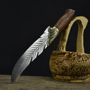An engraved feather blade knife displayed on a dark background, resting on driftwood next to a carved wooden basket.