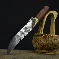 An engraved feather blade knife displayed on a dark background, resting on driftwood next to a carved wooden basket.