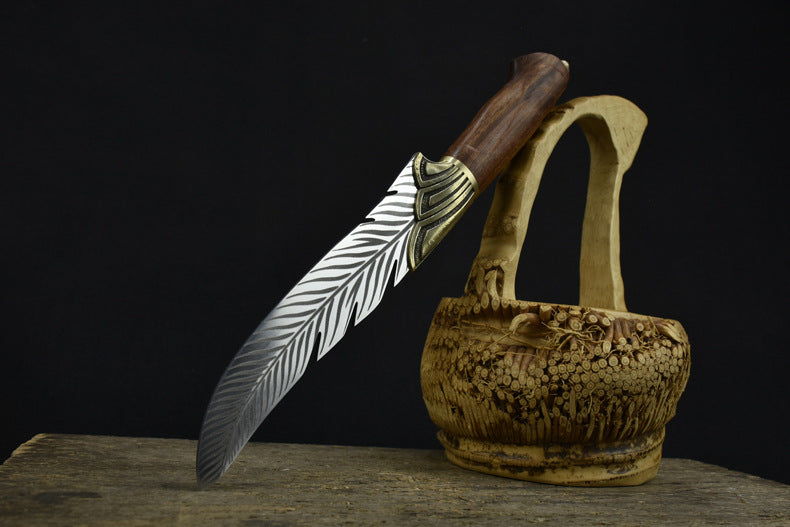 An engraved feather blade knife displayed on a dark background, resting on driftwood next to a carved wooden basket.