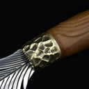 An extreme close-up of a custom fixed knife, highlighting the textured hammered brass bolster seamlessly connecting to the intricate silver feather-engraved blade, with a grooved wooden handle visible in the background.